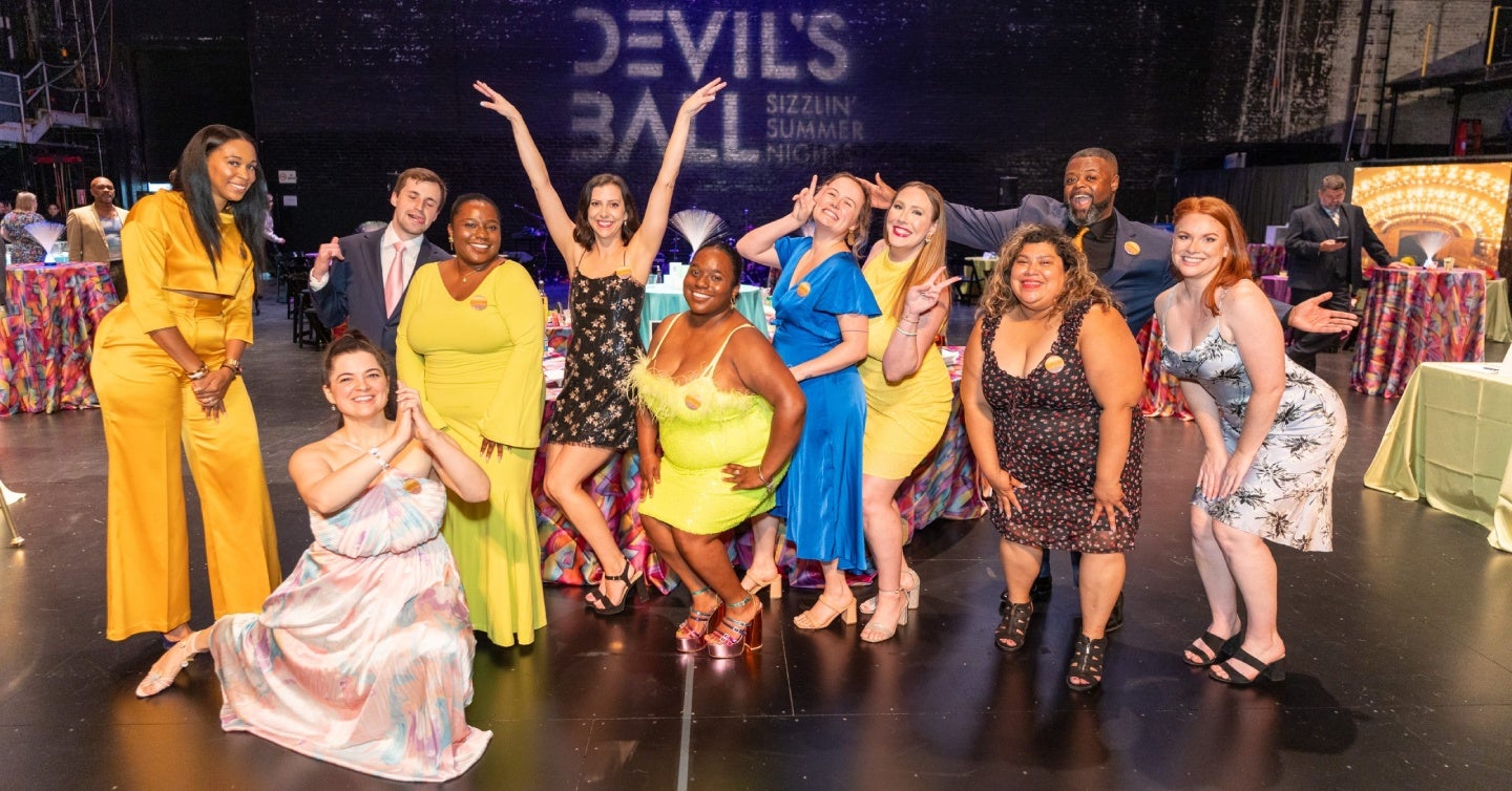 Group of people posing joyfully in colorful outfits at an event titled &quot;Devil's Ball,&quot; with a festive atmosphere and vibrant table settings in the background.