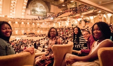 A group of five people sitting in an auditorium, smiling at the camera, with a large audience in the background.