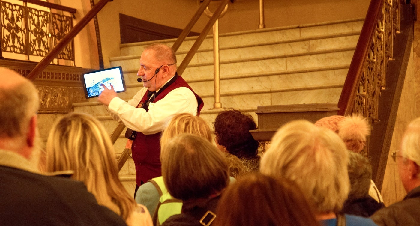 Tour guide speaking to a group while holding an image, standing on an ornate staircase.