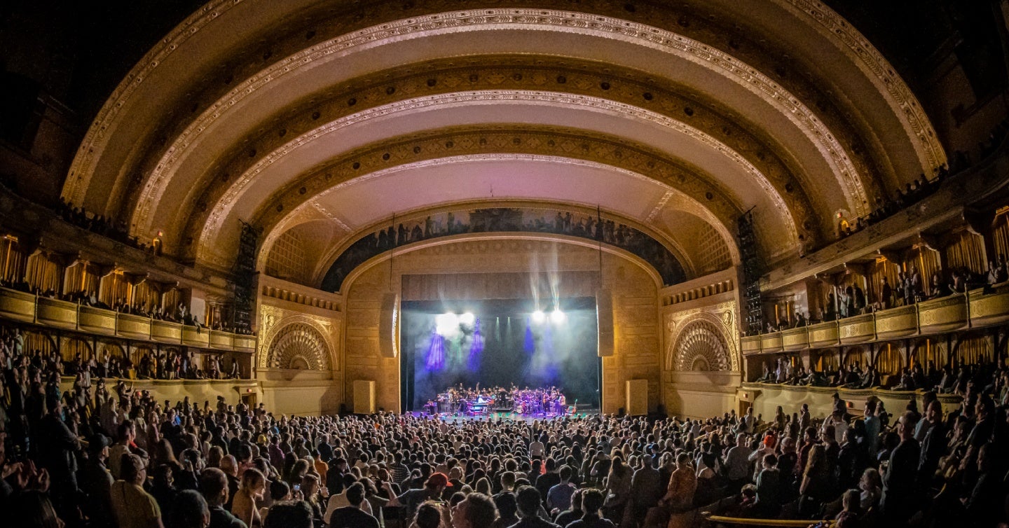 A large audience watches a live concert in an ornate, domed theater with illuminated stage lighting.